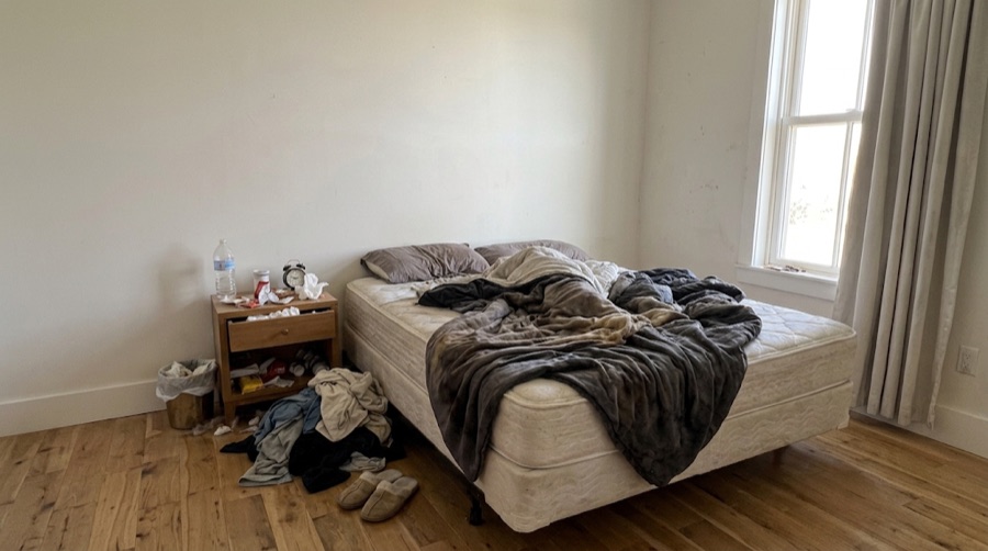 The bedroom before the turnover: the bed stripped back and unmade, clothes heaped on the floor, the bin overflowing, the nightstand cluttered.